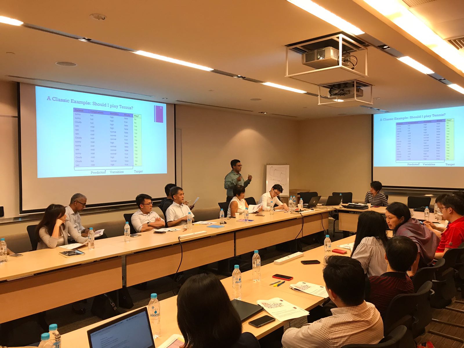 Workshop audience seated around tables facing a data table slide